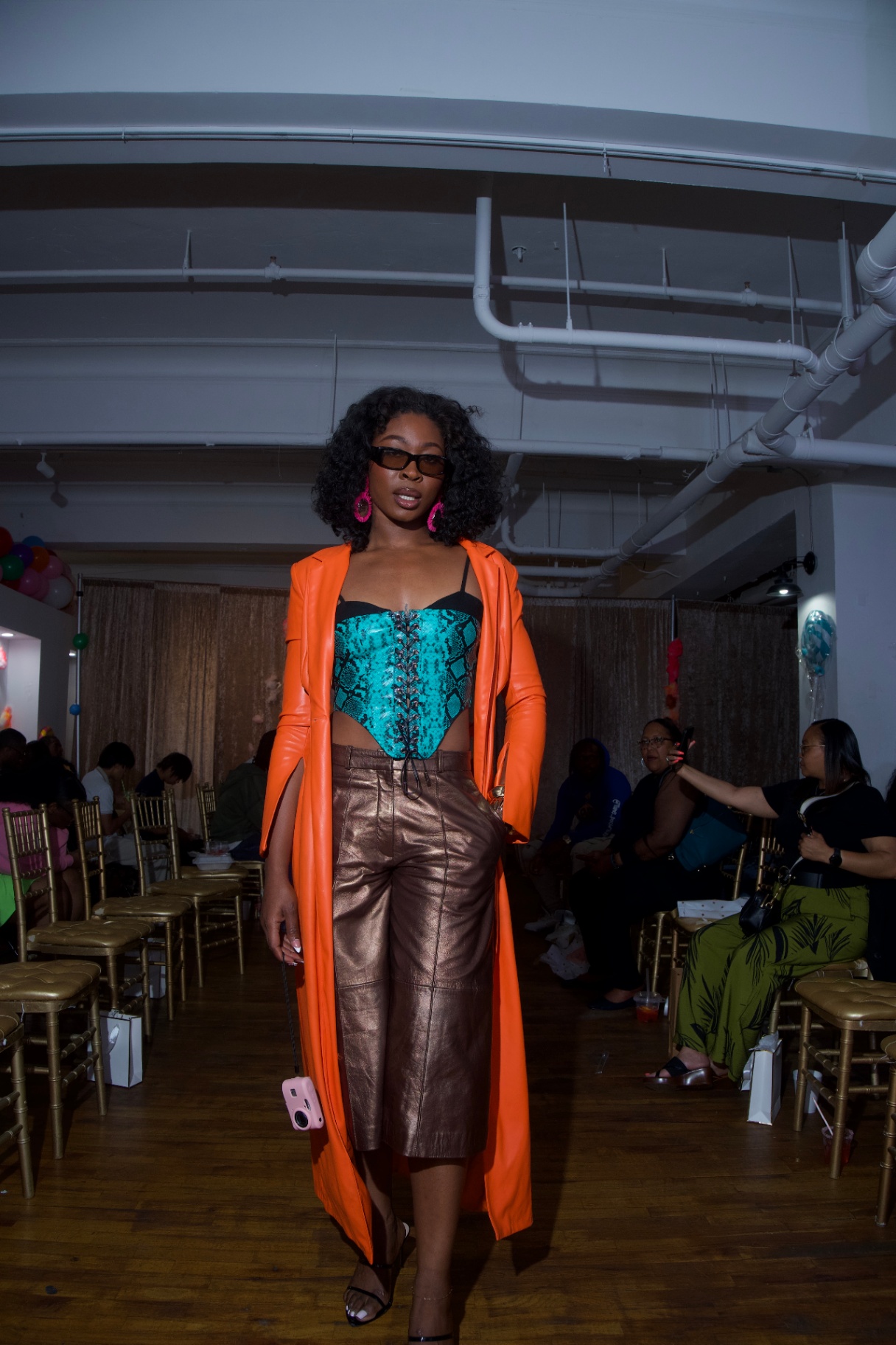 Guest in orange trench, teal snakeskin corset, and bronze leather pants